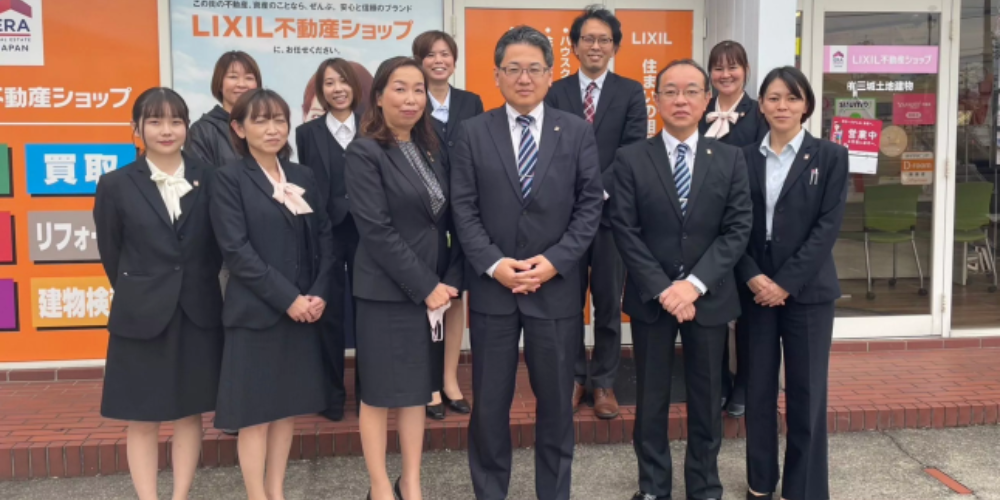 「LIXIL不動産ショップ」の店頭前でスーツ姿のスタッフ11名が横一列に並び、笑顔で記念撮影する集合写真
