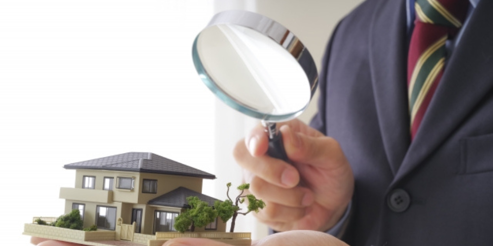 スーツ姿の人物が手のひらに乗せた住宅模型を虫眼鏡で精査している様子