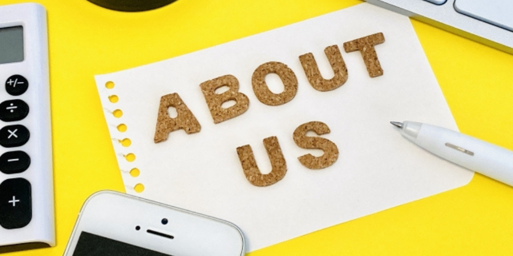 黄色い背景の上に「ABOUT US」と書かれた紙が置かれ、周囲に電卓、スマートフォン、ペン、キーボードが配置されている