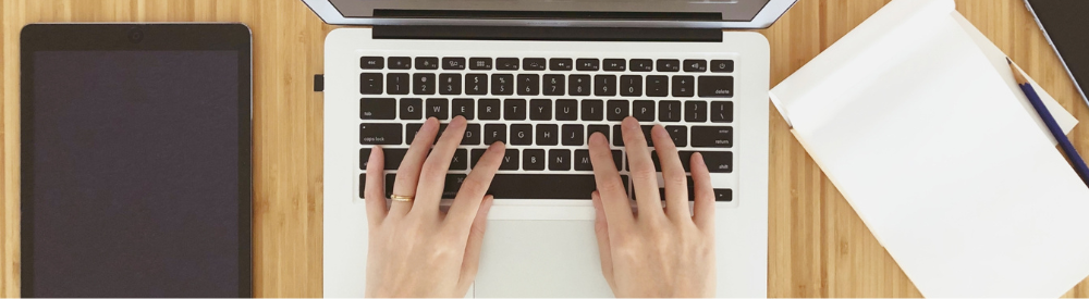 ノート PC をタイピングする両手を真上から撮影。左右にタブレットとメモ帳が置かれ、在宅で物件データを入力する作業イメージ