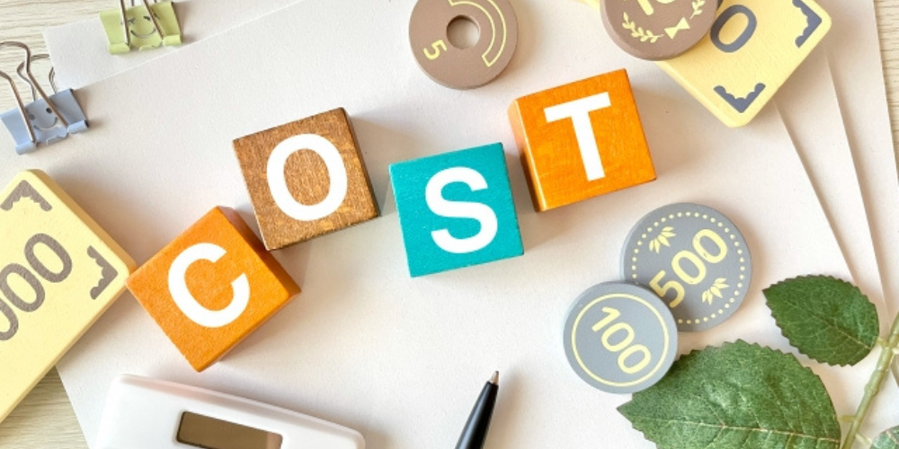 木製ブロックで「COST」と書かれ、周囲に紙幣デザインのパーツや電卓が置かれている画像