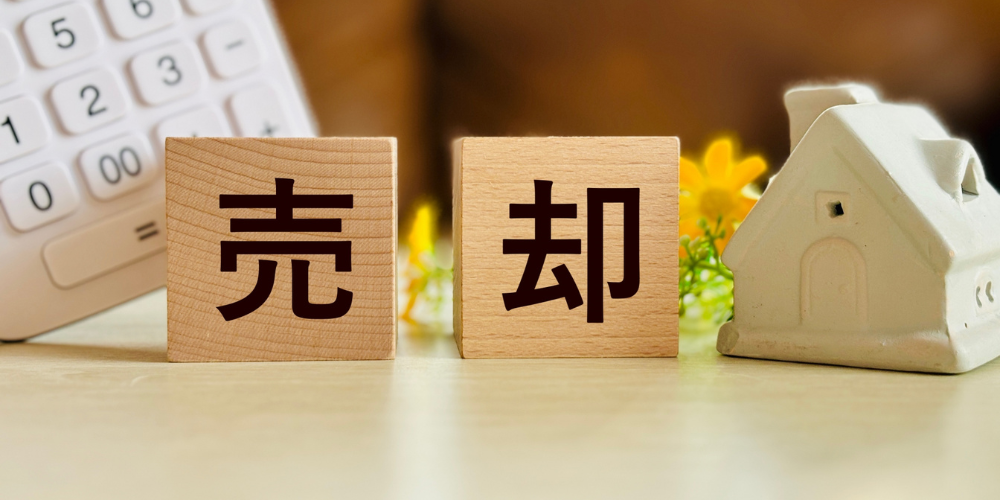 木製ブロックに「売却」と書かれた文字と小さな家の模型、背景には電卓と花