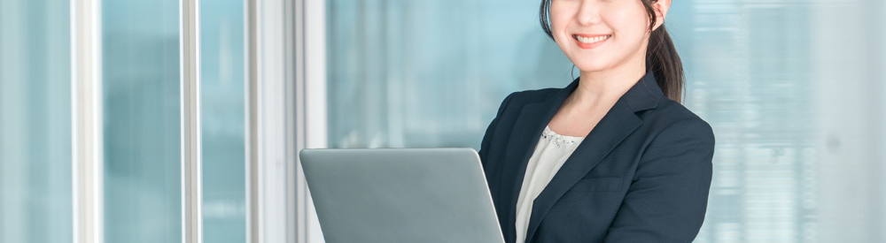 ガラス壁際でノートPCを抱え笑顔を向けるスーツ姿の女性営業。横長バナーサイズのビジネスシーン
