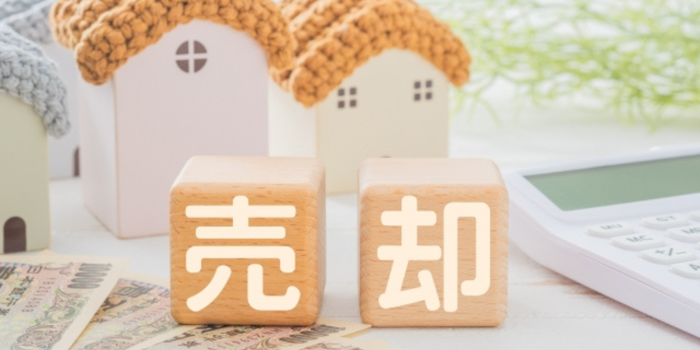 木製ブロックに「売却」と書かれ、家の模型や紙幣、電卓が周囲に置かれている画像