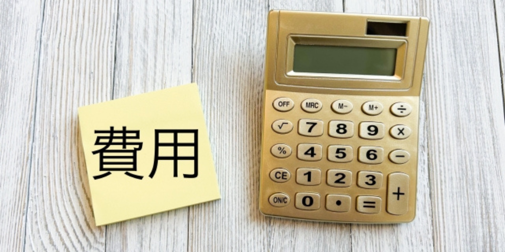 「費用」と書かれたメモとゴールドの電卓が木目の机の上に置かれている画像