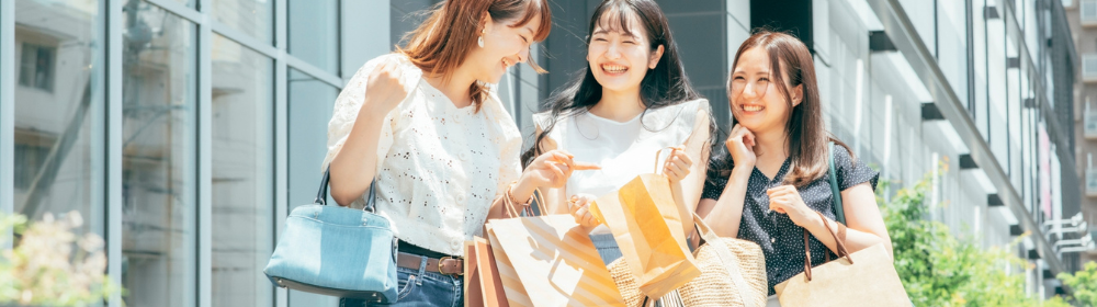 都市の商業施設前でショッピングバッグを手に笑顔で談笑する若い女性３人