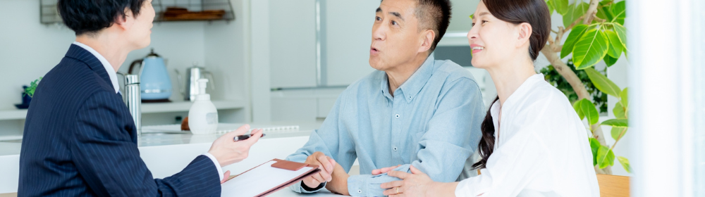リビングでスーツ姿の営業担当者と向かい合い、真剣に説明を聞く中高年夫婦の面談風景