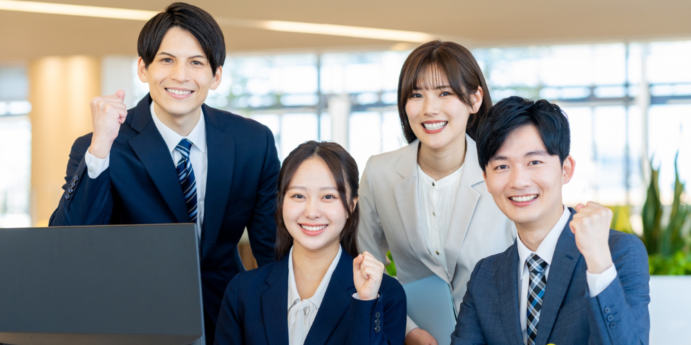 オフィス受付前でパソコンを囲み、スーツ姿の男女4人が拳を握り笑顔で意気込むチーム写真