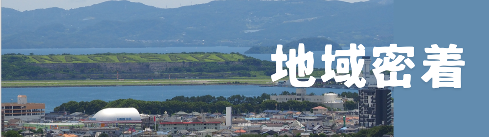 大村湾越しに「NAGASAKI」の植栽文字が見える長崎空港対岸の景観と市街地を俯瞰し、右側に白抜き文字で「地域密着」と掲げたバナー