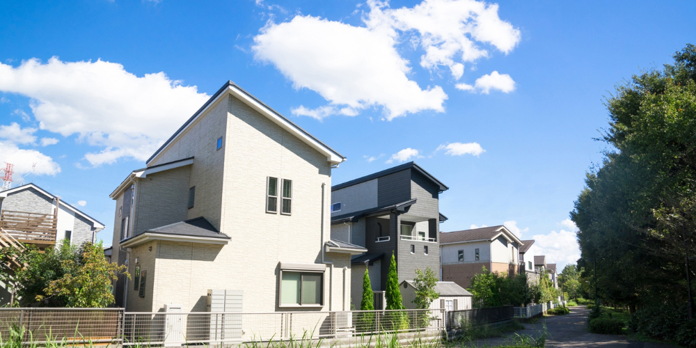澄んだ青空の下、緑豊かな通り沿いに建つ新興住宅街の戸建てが並ぶ街並みイメージ