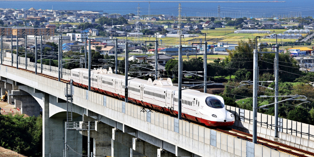 海と街並みを背景に、高架橋を滑走する白と赤の新幹線車両（かもめ）が走行する様子