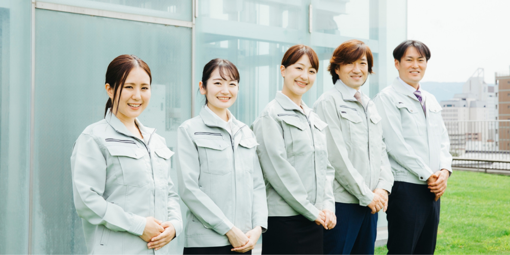 屋外で並んで立つ作業服姿の男女スタッフ５名、満面の笑みで会社をアピール